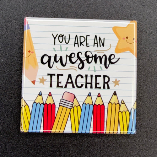 A square magnet featuring the text 'YOU ARE AN awesome TEACHER' on a white lined paper background, decorated with cartoon pencils and a smiling star, displayed on a dark textured surface.